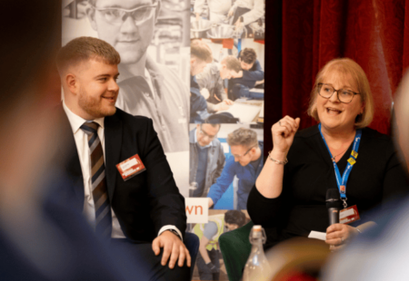 Professional female speaking on a panel on stage, professional male beside her and a image of apprentices behind.