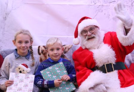 Two young children with wrapped gifts sat on the left of Santa, waving