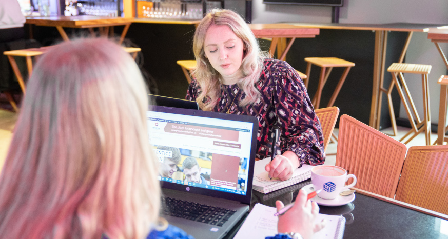 woman with blonde hair with a laptop sitting opposite another woman with blonde hair