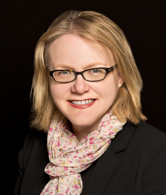 Woman with blonde hair and glasses smiling in front of black background