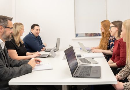 2 men and 4 women sitting round a white, rectangular table with laptops in front of them