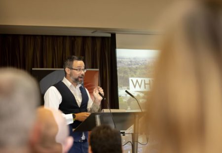 Man in waistcoats stood on a stage talking to people