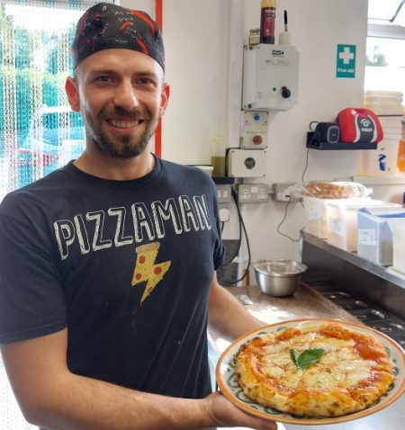 Chef wearing t shirt with pizza man written on the front holding a freshly cooked pizza in kitchen