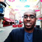 A male with glasses smiling on a street with unmbrellas hanging in the background - Ojay Mcdonald