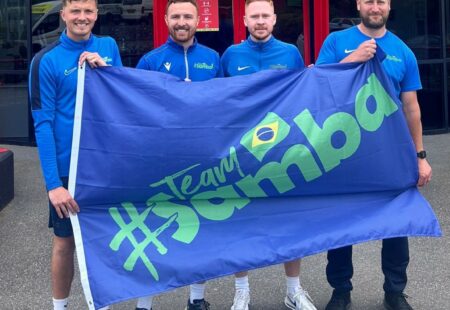 4 males holding a giant flag that reads #team samba - the 4 owners of samba sports coaching