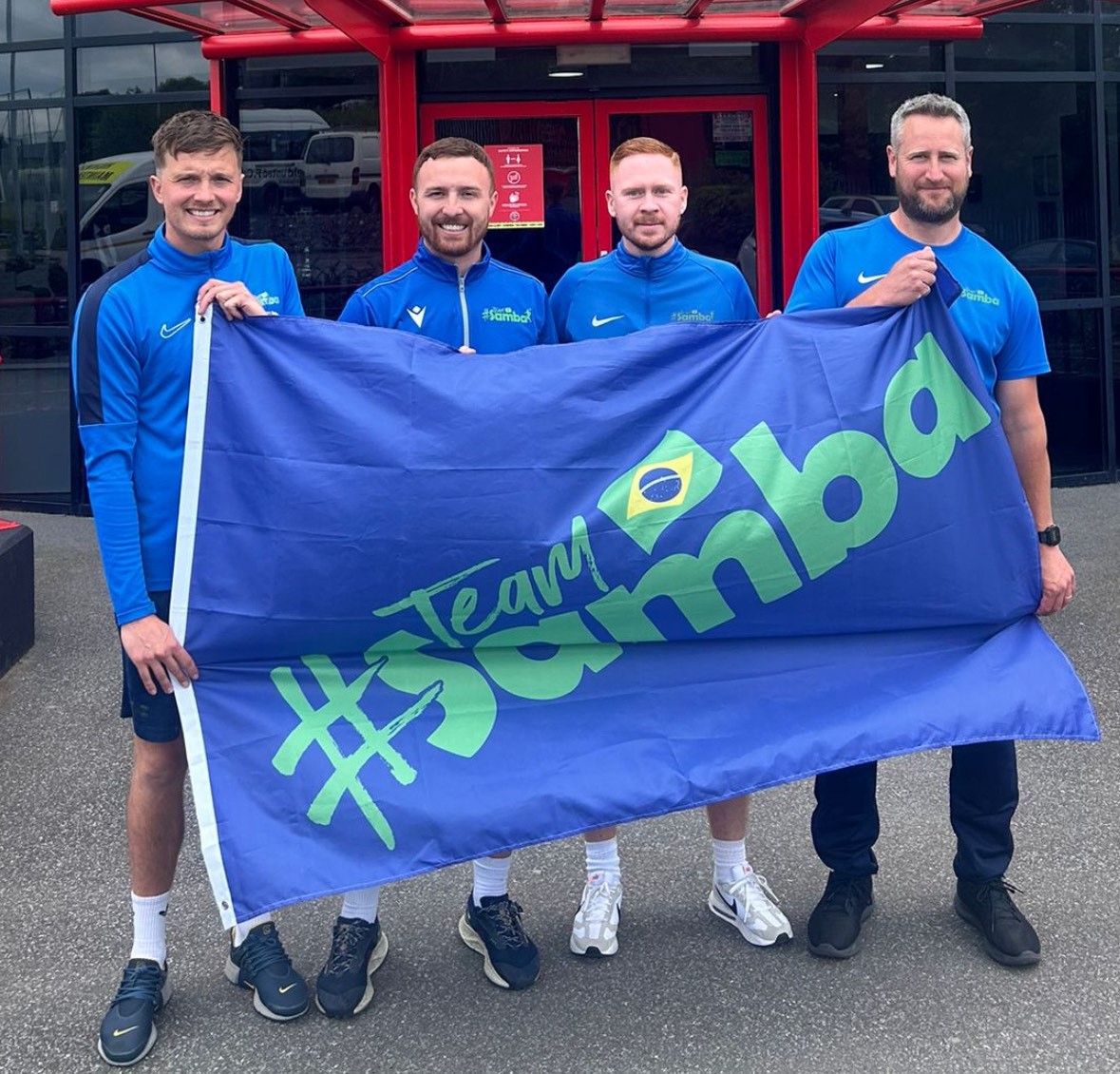 4 males holding a giant flag that reads #team samba - the 4 owners of samba sports coaching