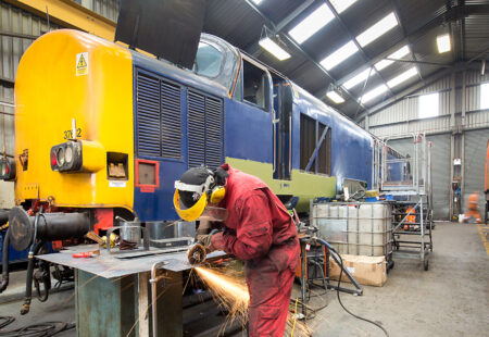 Mechanic welding metal with large rail locomotive in background