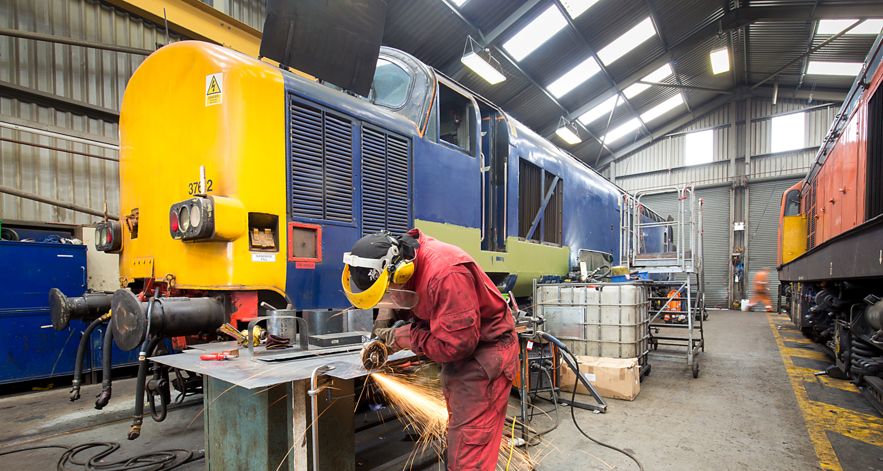 Mechanic welding metal with large rail locomotive in background