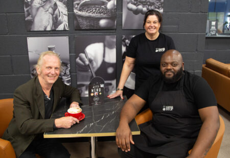 Three people smile for camera whilst gathered around table drinking a coffee