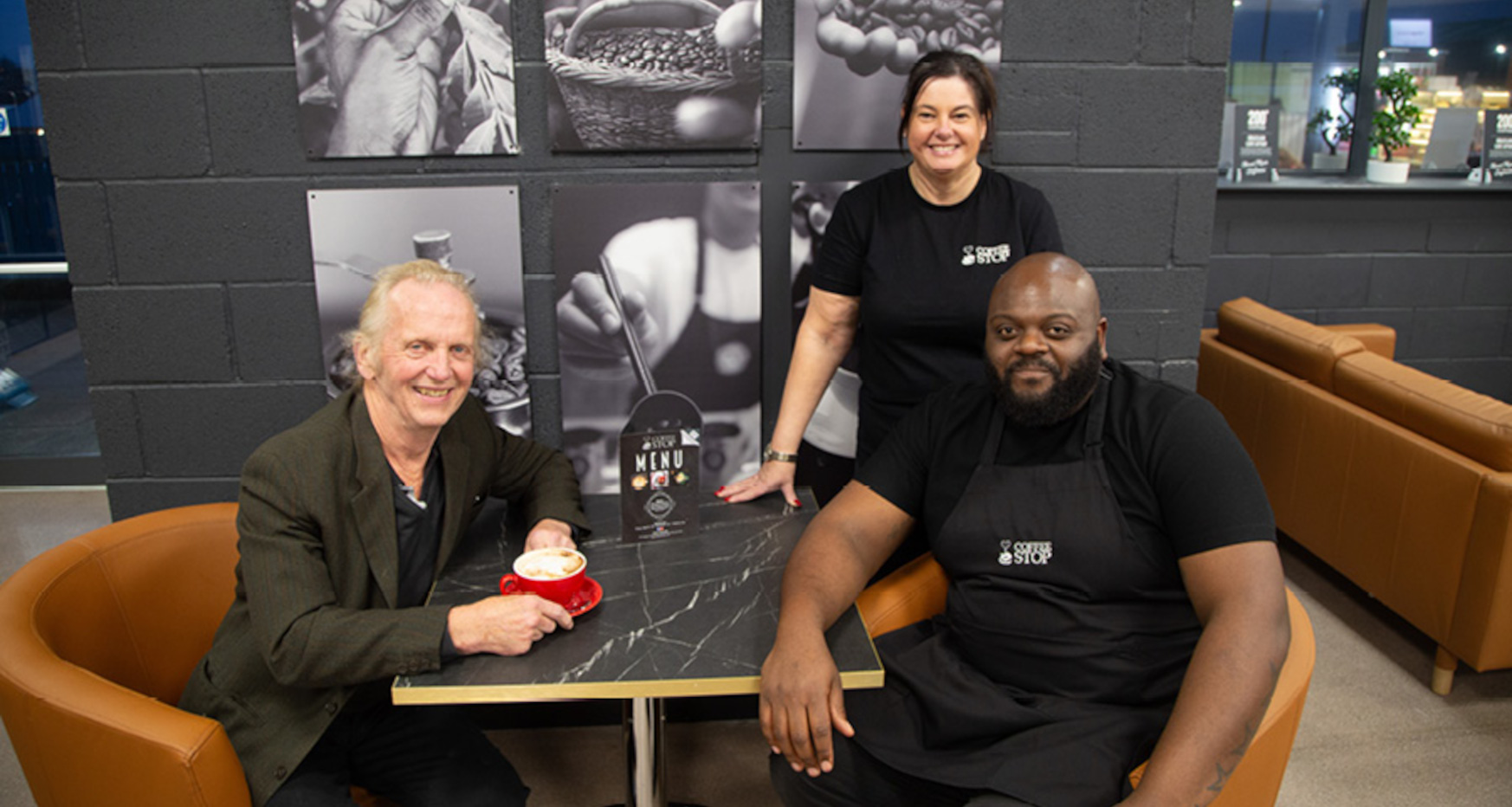 Three people smile for camera whilst gathered around table drinking a coffee